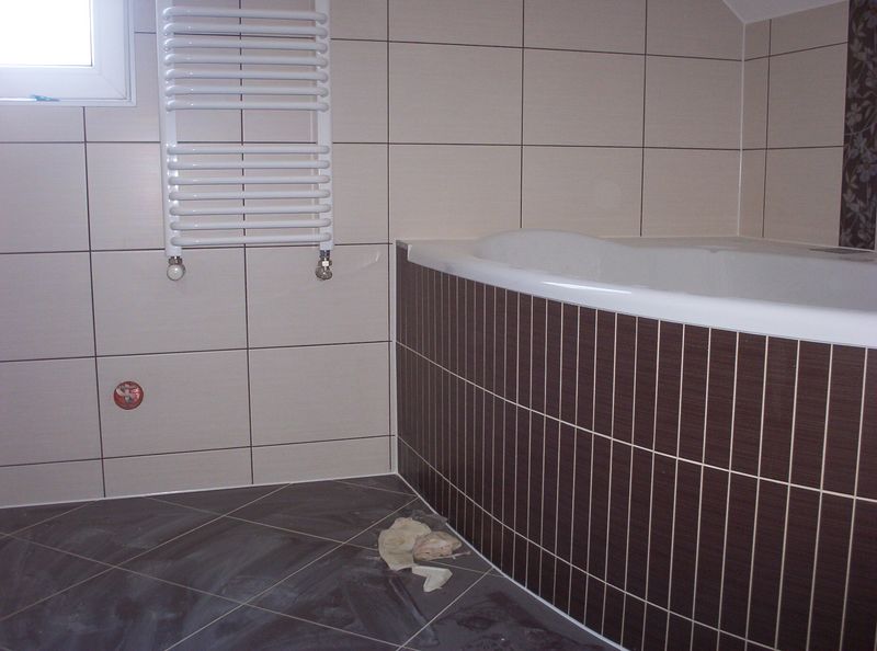 Wykończona łazienka z beżowymi płytkami ściennymi, ciemnymi płytkami podłogowymi ułożonymi ukośnie, oraz obudową wanny z brązowych płytek w pionowe paski; na ścianie biały grzejnik drabinkowy.